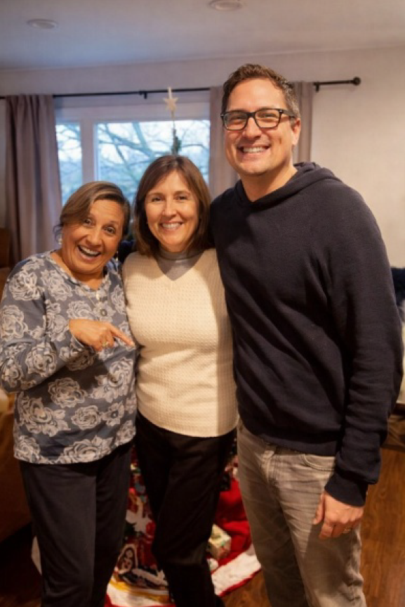 Three adults stand together indoors, smiling at the camera in a cozy living room setting.