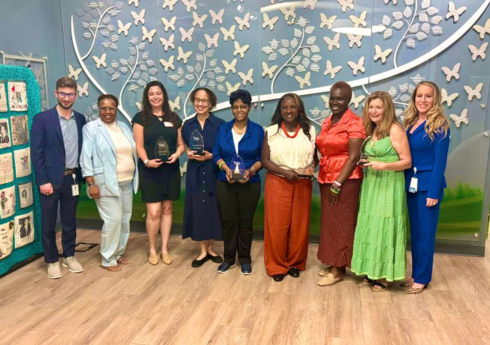 Nine professionals pose with glass trophies in a hallway with a blue butterfly mural behind them.