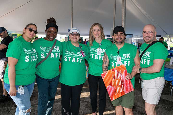 Group of seven event staff wearing green '5K STAFF' shirts posing together under a tent and smiling for the camera.