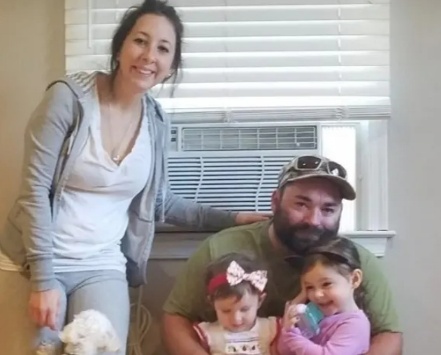 Smiling family of four posing indoors: a standing woman, a man with a hat, two young girls, and a small white dog nearby by the feet.
