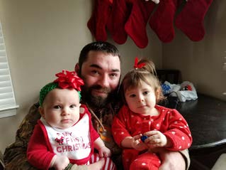 A man with a beard holding two small children in red outfits for a family photo indoors with holiday stockings overhead.