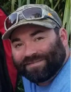 Bearded man wearing a camouflage cap and sunglasses on top, smiling outdoors.