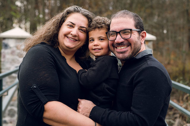 Adam-Bleznick-and-Familyforweb | NJ Sharing Network Smiling family of three hugging on a bridge outdoors; mother, father, and young child wearing black tops. A mom, dad, and child embrace and smile at the camera on an outdoor railing, autumn trees in background. Family portrait of parents holding their young child, all wearing black tops, posing on a park walkway. Close-knit family photo: mother on left, father on right, child in middle, smiling. Happy family moment on a bridge with trees in the background.