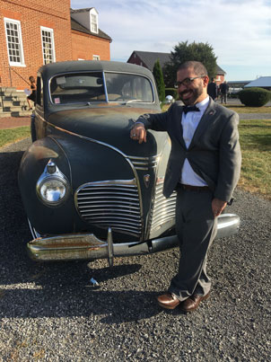 Adam-Bleznick1-for-web | NJ Sharing Network Man in a gray suit and bow tie stands beside a dark green vintage car on a gravel lot, with a brick building in the background.