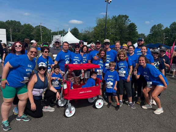 Adams-Warriorsforweb | NJ Sharing Network Group of people in blue race shirts posing for a photo at an outdoor race, centered around a red double stroller with kids in front.