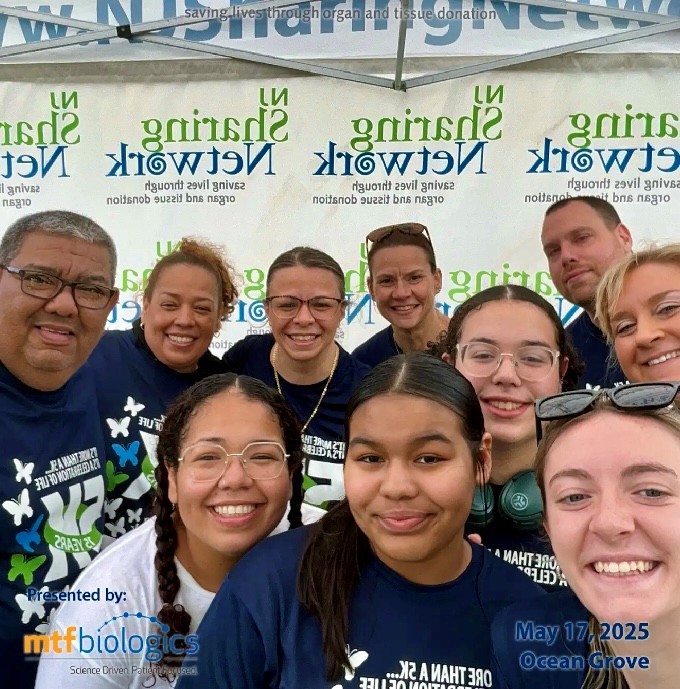 Karen Stash Oscar Salgado members of their 5K team | NJ Sharing Network Group of smiling volunteers in matching blue shirts posing for a group selfie at an outdoor outreach event, with banners and a 'Presented by mtfbiologics' decal visible.