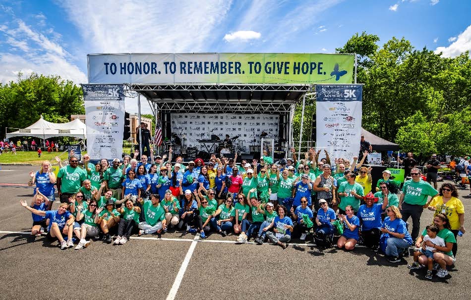 2026 New Providence 5K PR - Post Event | NJ Sharing Network Group photo of volunteers and participants in colorful shirts posing in front of a charity event stage with a banner: 'To Honor, To Remember, To Give Hope.'