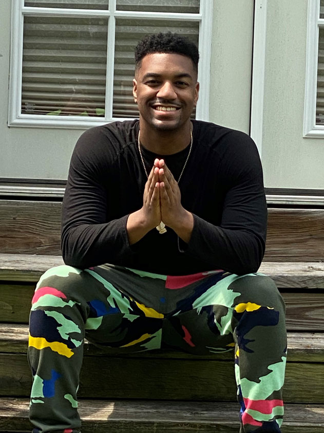 Jordan-Greene-for-web | NJ Sharing Network Smiling man sits on wooden steps with hands pressed together in a prayer pose, wearing a black long-sleeve shirt and camo pants.