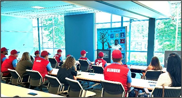 Group of people in red shirts and caps seated at desks in a classroom, facing a presenter beside a blue column with a poster board.