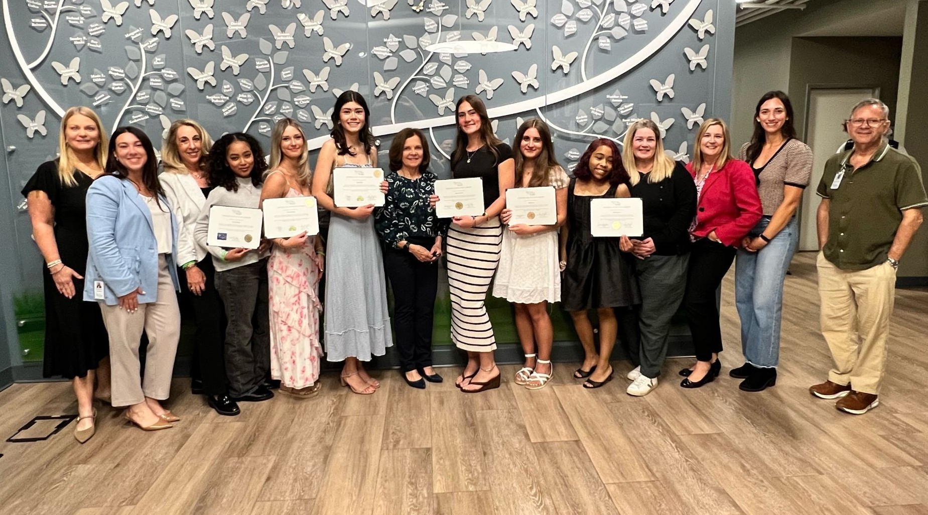 2026 High School Scholarship Winners Photo 1 | NJ Sharing Network Group of people smiling in a line indoors; several hold certificates, standing before a blue wall with white butterfly cutouts.