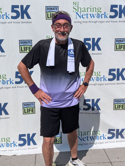 Chris-Carrales-1 | NJ Sharing Network Smiling man with gray beard and purple headband posing with hands on hips in front of a 5K event backdrop featuring Donate Life and NJ Sharing Network logos.