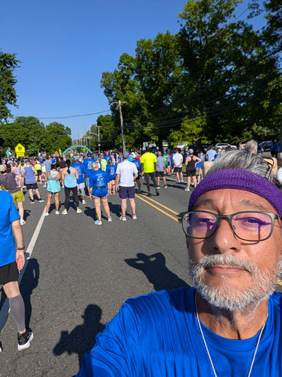 Chris-Carrales-3 | NJ Sharing Network Man in a blue shirt with a purple headband takes a selfie as runners line a sunny, tree-lined street for a road race.