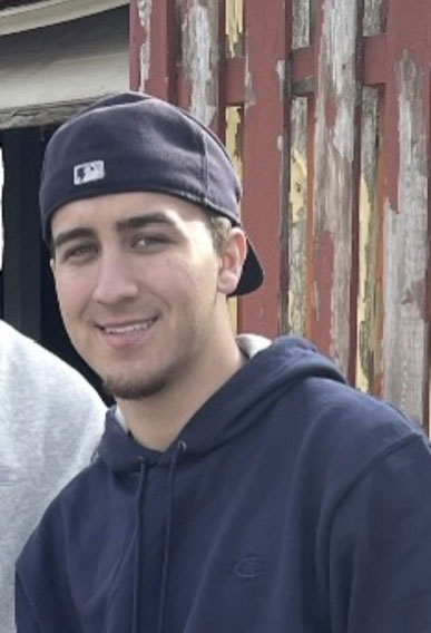 Gene-Patrick-Coppola-Jr | NJ Sharing Network Young man wearing a navy hoodie and backward baseball cap, smiling at the camera beside a weathered wooden fence.