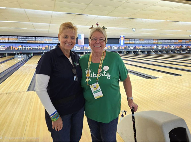 Two smiling women stand together at a bowling alley; the woman on the left wears a dark polo and the woman on the right wears a green 'Liberty' shirt with a lanyard and badge, and she holds a cane while the bowling lanes stretch behind them.