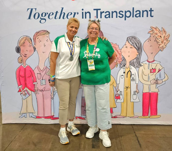 Two smiling women posing at a transplant event in front of a 'Together in Transplant' banner, wearing lanyards and medals.