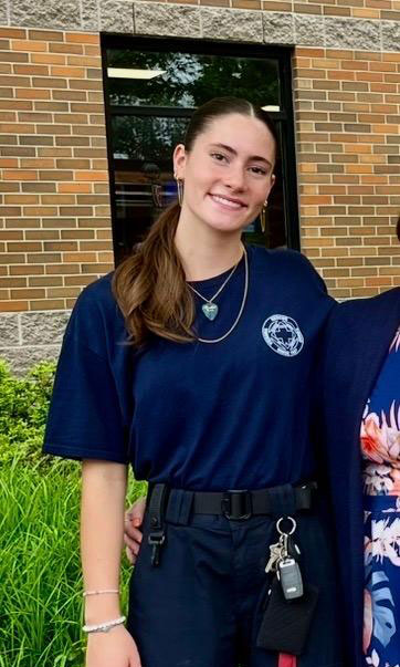 Jessica-Theobald | NJ Sharing Network Young woman with long brown hair in a navy uniform, smiling outside a brick building with keys clipped to her belt.