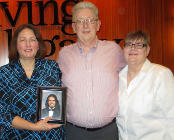 Three people stand together; the woman on the left holds a framed photo of a bearded man, with a smiling man in the middle and a woman on the right.