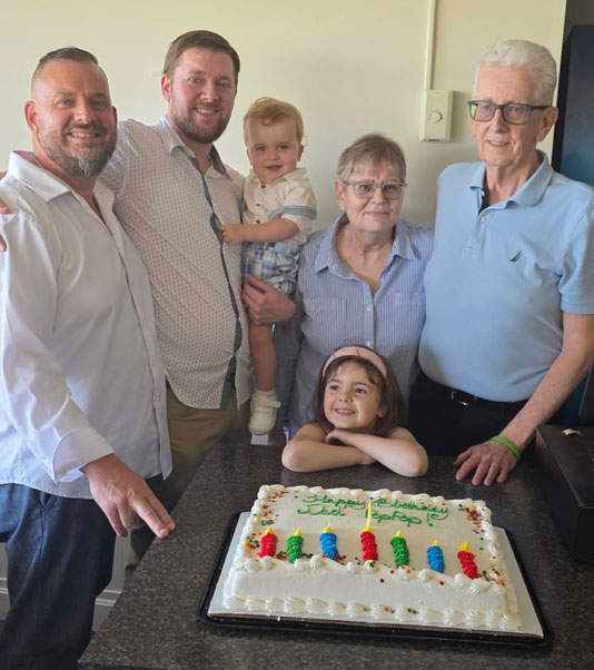Multigenerational family gathered around a birthday cake on a kitchen counter, smiling at the camera with a young girl resting her chin on the cake plate edge.