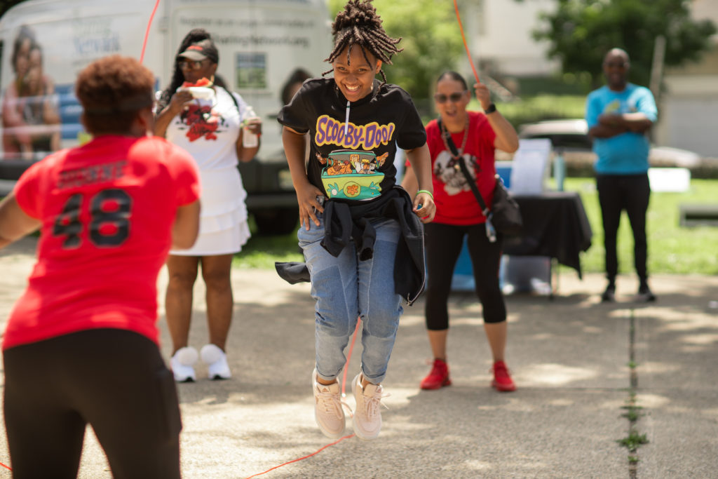 LHM-133 Girl in a Scooby-Doo shirt jumps rope while others watch at an outdoor event on a sunny day.