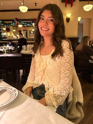 Leyla-Rolon | NJ Sharing Network Woman with long dark hair wearing a cream lace blouse smiles at the camera while seated at a festively decorated restaurant table.