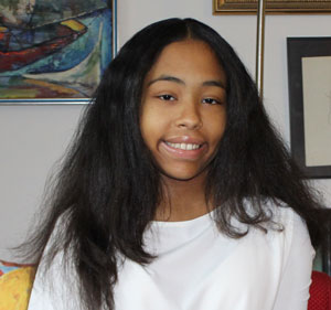 Olivia-Huttinot | NJ Sharing Network Young woman with long dark hair and a white shirt, smiling at the camera indoors with framed artwork in the background.