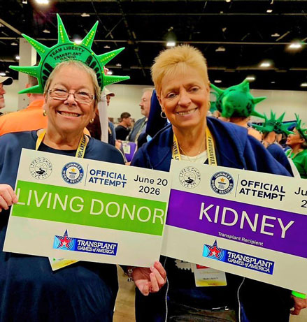 Two smiling women at a Transplant Games event wear green Statue of Liberty hats and hold 'LIVING DONOR' and 'KIDNEY' signs.