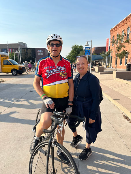 Rich-and-Donna-Albanese-DeMair-1 | NJ Sharing Network Man in red-yellow cycling jersey and helmet posing with a smile beside a woman on a sunny city street with a road bike in front of them.