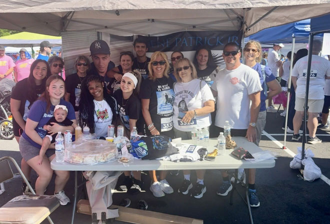 Team---Gene-Strong-1 | NJ Sharing Network Diverse group of adults (and a baby) pose behind a table with snacks and promotional items at an outdoor charity event under a white canopy.