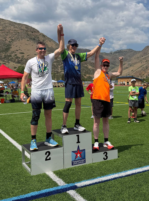 Three male runners stand on a winner's podium (1st, 2nd, 3rd) raising their arms in victory on a football field with mountains in the background, medals around their necks.