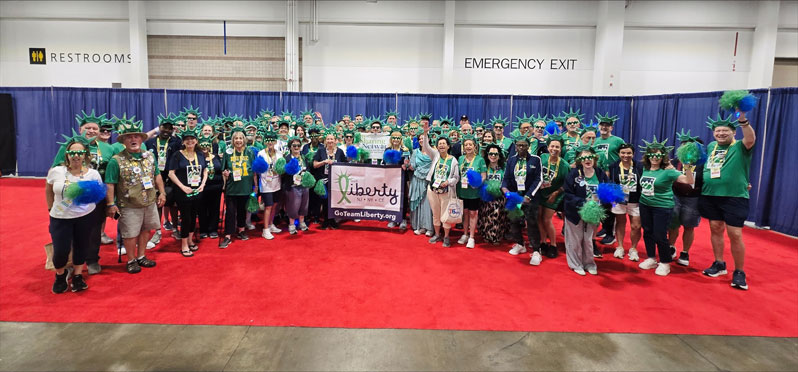 Large group of people wearing green shirts and Statue of Liberty hats posing on a red carpet, holding a Liberty-themed banner in a convention hall.