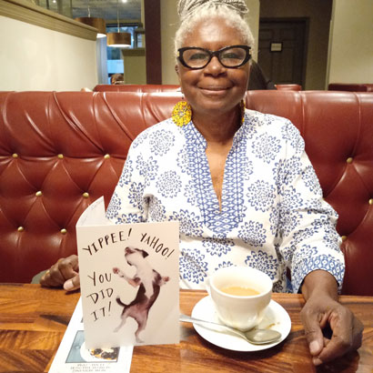 Gloria-Robinson-Mills-1 | NJ Sharing Network Smiling older woman in a blue floral blouse sits in a red leather booth with a celebratory card and a cup of coffee at the table.