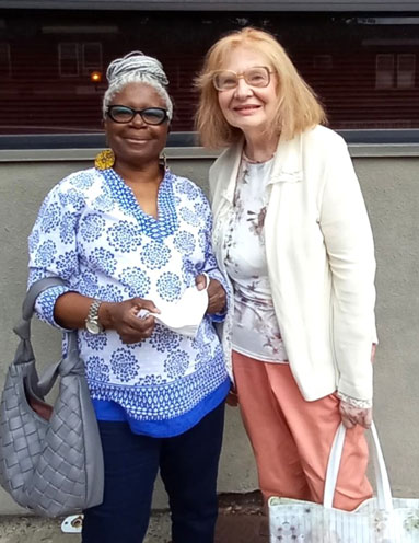 Gloria-Robinson-Mills-and-her-friend-Denise-Cooney | NJ Sharing Network Two women posing outside a building; left is a Black woman in a blue patterned blouse with a gray bag, right is a white woman in a cream cardigan and peach pants, holding a white face mask.