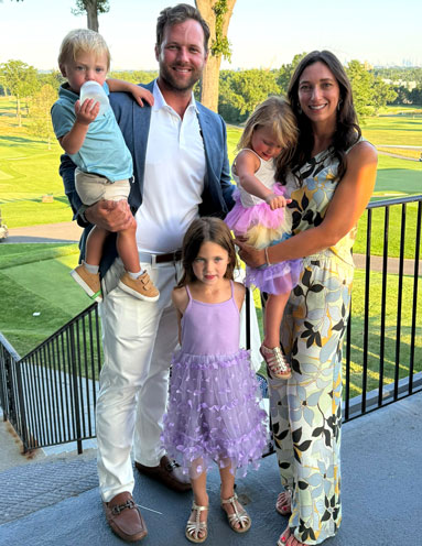 John-Levine-Fund-Golf-Event-1 | NJ Sharing Network Family of five posing on a balcony at a park: dad in a blazer with a son, mom in a floral dress with a toddler, and two daughters in lavender dresses.