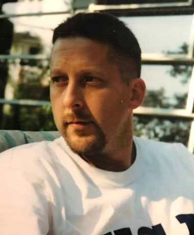 John-Levine | NJ Sharing Network Portrait of a man with short dark hair and a goatee, wearing a white T-shirt, outdoors near glass windows.