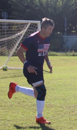 1---Matt-Felix-of-the-US-Transplant-Soccer-Team | NJ Sharing Network Futbolista con una camiseta con la bandera estadounidense, calcetines blancos, botas rojas y una rodillera, corriendo por un campo de césped cerca de la portería.