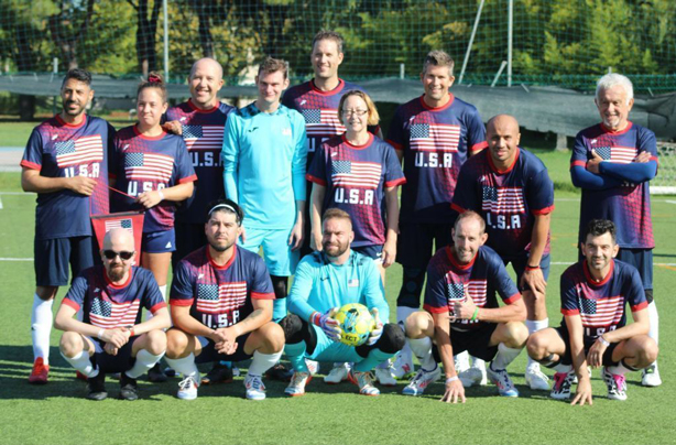 2---US-Transplant-Soccer-Team | NJ Sharing Network Grupo de futbolistas estadounidenses posando para una foto de equipo en un campo verde al aire libre; camisetas de color rojo marino con la bandera de EE. UU.; el portero, vestido de azul claro, está arrodillado en el centro, en primera línea, sosteniendo un balón.