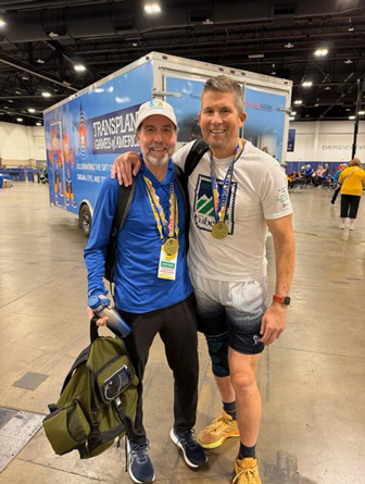 3---Patrick-Kacani-and-Matt-Felix | NJ Sharing Network Dos hombres con medallas posando juntos en un evento de los Transplant Games America celebrado en un recinto cubierto, con una pancarta azul del evento detrás de ellos.