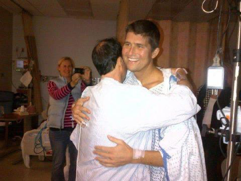 4---Matt-Felix-embraces-Patrick-Kacani-following-transplant-in-2012 | NJ Sharing Network Dos hombres se abrazan en una habitación de hospital; uno de ellos lleva una bata de hospital, mientras que una mujer, al fondo, les hace una foto.