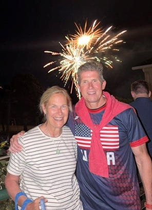 6---Matt-and-Trish-Felix-1 | NJ Sharing Network Dos adultos sonriendo por la noche con fuegos artificiales estallando a sus espaldas; una mujer con una camiseta a rayas y un hombre con una camiseta de la selección de EE. UU. y una bufanda roja.