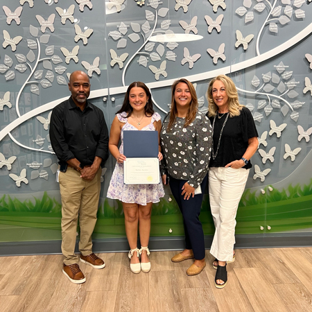 Amber-Astalos_presentation | NJ Sharing Network Young girl in a lavender dress holding a certificate, flanked by three adults, in front of a butterfly-patterned wall.