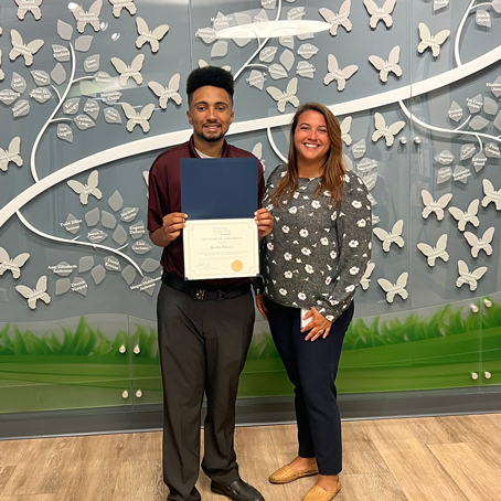 Andre-Silvera_presentation | NJ Sharing Network Man in a maroon shirt holds a blue-covered certificate as he stands with a smiling woman in a floral sweater; butterfly-themed wall and green mural behind them.
