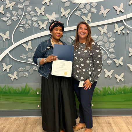 Clirver-Farrow_presentation | NJ Sharing Network Two women stand side by side smiling; the left holds a certificate in a blue folder, in front of a wall covered with white butterfly decorations.