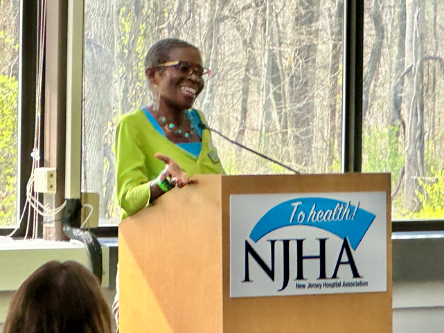 E-Denise-Peoples-2 | NJ Sharing Network Smiling woman in a bright green cardigan speaks at a podium with an NJHA banner in front of a windowed backdrop sports a turquoise necklace and glasses.