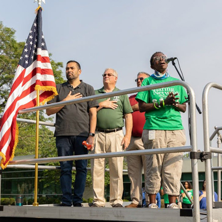 E-Denise-Peoples-3 | NJ Sharing Network Diverse group on a stage with an American flag; a man at a microphone speaks while others place hands over their hearts outdoors.