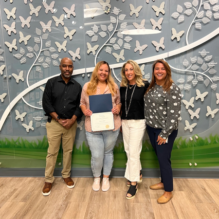 Erica-Hall_presentation | NJ Sharing Network Four adults stand indoors; a woman second from left holds a certificate in front of a butterfly mural.