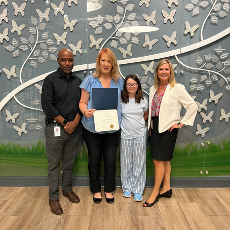 Katherine-Marino | NJ Sharing Network Four people pose for a photo in front of a butterfly wall; woman center holds a certificate.