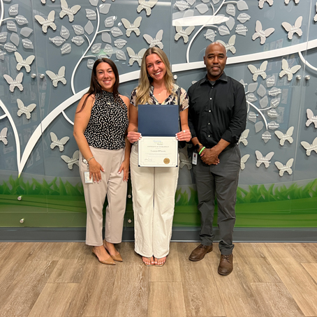 Lauren-OLeary_presentation | NJ Sharing Network Center woman holds a diploma in a blue folder, flanked by two colleagues, posing in front of a butterfly mural and wooden floor.