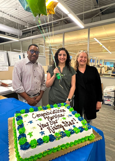 Three coworkers pose around a decorated cake on a blue table, with green/blue frosting and balloons overhead; the center woman holds green, blue, and yellow balloons.