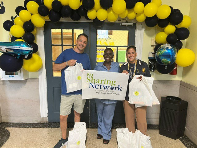 Tres voluntarios sonrientes sostienen una pancarta de NJ Sharing Network en el umbral de una puerta, rodeados por un arco de globos amarillos y negros, con bolsas para donaciones cerca.