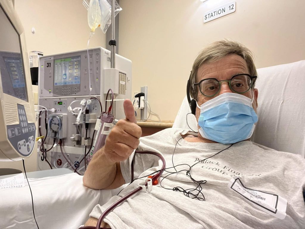 Patient in a hospital bed wearing a blue mask and glasses, giving a thumbs-up with medical equipment (dialysis machines) in the background labeled Station 12.